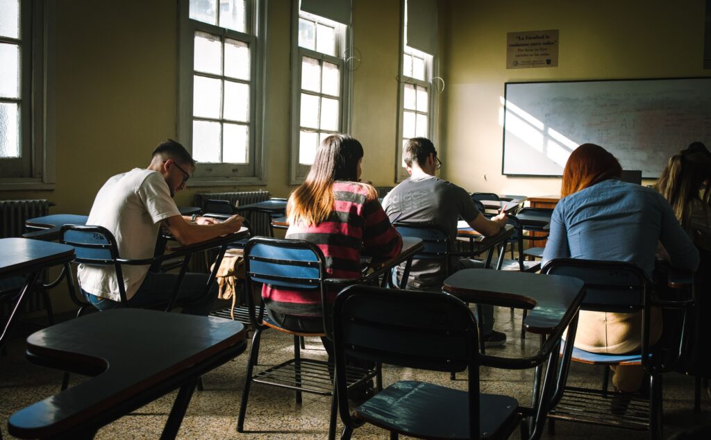 A group of students taking an exam in a classroom, illuminated by natural sunlight from windows.